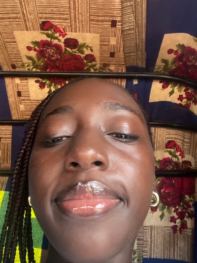 A close-up selfie of a young Black woman is captured from a low angle. She has braided hair and is wearing a gold hoop earring. Her face is illuminated by bright lighting, highlighting her glossy lips and smooth skin. The background features a patterned fabric with floral motifs in shades of red and cream, interspersed with brown stripes and blue panels. The image appears to have been taken indoors. There is no specific indication of the location being Galadimawa, Nigeria.