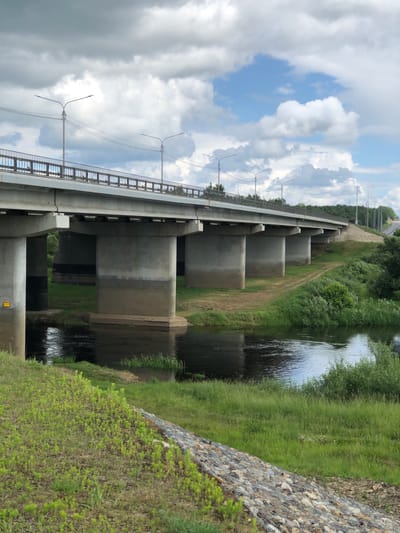 The image shows a concrete bridge spanning a river. The bridge has multiple piers supporting the deck, which is lined with railings and streetlights. The river flows beneath the bridge, with lush green vegetation growing along its banks. The sky is partly cloudy with a mix of blue sky and white clouds. A dirt path leads up to the bridge on the right side of the image. The location is identified as Nadezhda, Russia. There are no people visible in the image. The scene depicts a typical rural or suburban landscape with a significant infrastructure element.