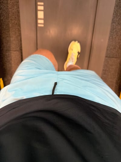 A top-down view depicts a person from the mid-torso down, engaged in an activity on a treadmill. The individual wears light blue athletic shorts over a black undergarment. The right leg is visible, with a foot clad in a bright yellow athletic shoe featuring black details and a white sole. The left leg and foot are partially obscured and blurred, consistent with forward motion. The treadmill belt is dark gray, with faint parallel light-colored markings. Dark gray side rails flank the belt. The background elements are out of focus and dark. The specific location within Munggu, Indonesia, cannot be determined from the visual information presented in the image.