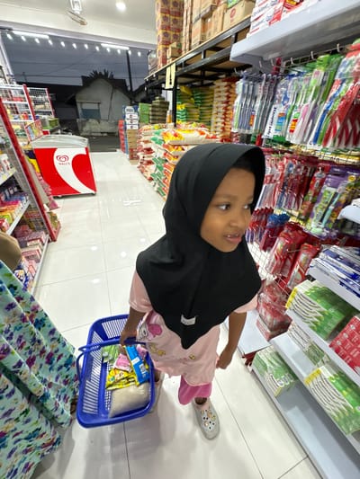 This is a slightly tilted, eye-level shot of a young girl in a convenience store in Kebonsari, Indonesia. The girl is wearing a black hijab and a light pink shirt with cartoon characters. She is holding a blue plastic basket filled with groceries, including what appear to be snacks and a bag of rice. Her left hand is gripping the basket handle, and her right hand is reaching for items inside. She is looking up and to the right with a slight smile, suggesting engagement or curiosity.
The background shows rows of shelves stocked with various products, primarily toiletries like toothbrushes and toothpaste, evident from the visible text such as "Ciptadent," "Pepsodent," and "Systema." There are also bags of rice and other packaged goods. A red and white "Wall's" refrigerator for ice cream is visible on the right side of the frame. The store appears well-lit with fluorescent lighting, and the floor is tiled. The scene is indoors, likely during the day, given the brightness. The overall impression is a candid moment of a child shopping with her family, interacting with the products around her.
