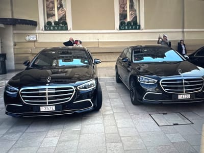 Two black Mercedes-Benz S-Class sedans are parked side-by-side on a tiled pedestrian walkway. The car on the left has license plate "357E" and the car on the right has license plate "432E". In the background, a building with large display windows for "GRAFF" jewelry is visible. Several people are in front of the building, including one individual taking a photograph with a cell phone. There is a sign pointing to an elevator. The scene appears to be in an urban environment, likely a commercial or luxury district.