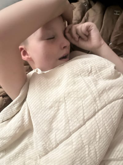 The image shows a close-up of a young person, likely a child, sleeping in bed. The person is lying on their side with their eyes closed and their hand resting on their forehead. They are covered with a light-colored, textured blanket. The background appears to be a bed with brown bedding. The image is taken from a slightly elevated angle. The scene is indoors and appears to be a domestic setting. There is no visible indication of the city of Peremyshliany, Ukraine.