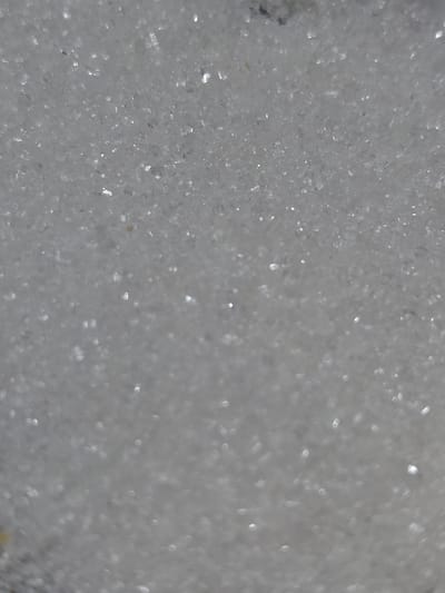 The image displays a macro photograph of fine, granular white material, likely sugar or salt crystals. The lighting creates numerous bright highlights and some bokeh effects, indicating a close-up perspective. The texture is rough and uneven due to the individual particles. There is no discernible environment or location evident in the image, and no people, subjects, objects, or actions are depicted. Therefore, it is not possible to determine the location in Ертіс ауылдық округі, Kazakhstan.