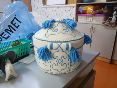 In a room, a felt-covered box with blue trim and tassels sits on a gray desk. Behind it, a blue plastic bag with "SEMET" printed in black lies on a green plastic basket containing a potted plant. A small felt animal figure is also on the desk. In the background, shelves hold snacks and containers, and a cabinet with silver handles is visible. The orange floor suggests an indoor setting, possibly a home or workshop in Taraz, Kazakhstan.