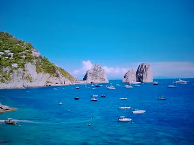 This is a wide-angle, high-angle shot of a picturesque coastal landscape under a clear blue sky. In the foreground, a calm, deep blue sea is dotted with numerous boats of various sizes, including speedboats, yachts, and smaller vessels. The water exhibits a gradient of blues, becoming lighter and more turquoise closer to the shore. On the left side of the image, a steep, rocky cliff covered in lush green vegetation rises dramatically from the sea. Several buildings, appearing to be houses or villas, are nestled into the hillside, characteristic of a Mediterranean coastal town. In the middle ground, two imposing rock formations, known as "Faraglioni" or sea stacks, rise majestically from the water. More boats are anchored around these iconic formations. The background is a vast expanse of clear blue sky with a few wispy white clouds. The scene evokes a sense of leisure, natural beauty, and vacation.
This scene is not located in Huntingdonshire, United Kingdom, but rather depicts the iconic coastline of Capri, Italy, known for its dramatic cliffs and the Faraglioni rock formations.