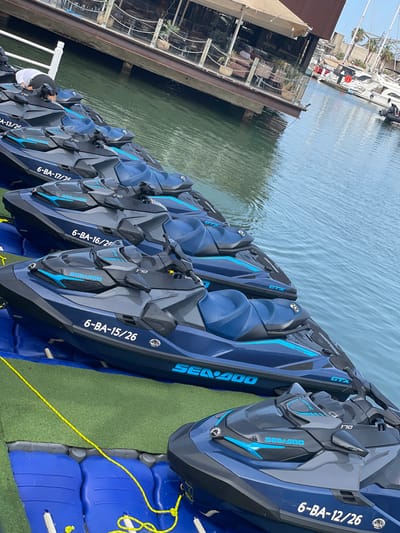 This image depicts a row of navy blue Sea-Doo jet skis docked on a waterway. The jet skis are lined up next to a floating dock covered in green astroturf, with blue plastic pontoons visible underneath. A person wearing a white shirt and a dark hat is partially visible, standing on the dock near the jet skis.
Several jet skis have visible text, including "ROTAX", "GTX", and "SEA-DOO", along with alphanumeric identifiers such as "6-BA-17/25", "6-BA-16/2", "6-BA-15/26", and "6-BA-12/26". The background shows a body of water with ripples, suggesting a calm day. In the upper left corner, there are glimpses of boats and buildings, hinting at a marina or waterfront area. The lighting suggests it is daytime. The overall scene is one of organized watercraft ready for use or rental in a sunny, possibly coastal, environment.