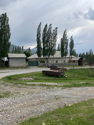 The image depicts a rural scene in Жаңы-Коргон, Kyrgyzstan, likely on an overcast day. Tall, slender trees with dense foliage line the left side of a gravel road that cuts through a grassy area. In the background, a long, two-story building with a white, corrugated metal roof stands prominently. The building has several windows and appears to be a residential or utility structure.
Adjacent to the building, a dark green metal gate is visible, suggesting an entrance to a property. In front of the building, a grey car is parked next to a rustic trailer or flatbed truck loaded with metal sheeting. Further down the road, another smaller, concrete building with a corrugated metal roof is situated.
The landscape is characterized by rolling hills or mountains in the distance, partially obscured by clouds and mist, hinting at the mountainous terrain of Kyrgyzstan. The grass on either side of the road is green and lush, with some scattered yellow wildflowers visible. The overall atmosphere is one of quiet rural life. There is no visible text in the image.
