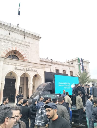 A crowd gathers outside a building with Syrian flags displayed. The building has arched windows and detailed stonework. A large truck with a digital screen displaying “Ministry of Sports and Youth” in Arabic and English is parked nearby. Men in various attire, including casual wear and uniforms, stand in front of the building. Security personnel with rifles are visible inside the building's entrance. The sky is overcast.