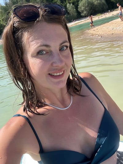 A woman with brown hair and blue eyes smiles at the camera. She is wearing a dark green bikini and sunglasses pushed up on her head. The woman is in the water, which appears to be a lake or river. In the background, there are trees and a sandy beach with several people. The location is the Lac de Vouglans in Maisod, France. The lighting suggests it is a sunny day.