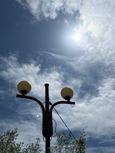 The image depicts a low-angle view of a street lamp against a partly cloudy sky in Ogradna, Bulgaria. The sun is visible in the upper left corner, partially obscured by thin clouds. The street lamp has two spherical, yellow lights attached to a brown, rusty metal structure. A tree with green foliage is visible on the right side of the frame, with an electrical wire running near its branches. The sky is a mix of blue and white, with cirrus clouds scattered throughout.