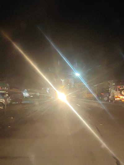 In Dattapur, India, at night, a street scene is captured with strong light flares from oncoming vehicles obscuring much of the detail. A white van is visible in the center-left, appearing to be stationary or moving slowly. To the right of the van, there is a dark-colored SUV, possibly a Jeep, also stationary or moving slowly. Several other vehicles are present in the background, some appearing to be trucks, but their features are indistinct due to the lighting conditions. The environment appears to be a road with some debris or uneven surfaces. The lighting suggests a bright light source, possibly headlights from a vehicle, is directed towards the camera, creating lens flares and washing out much of the image. The overall scene depicts a nighttime road with traffic, characterized by intense light and limited visibility of the surroundings.