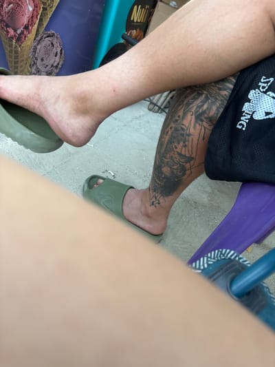This outdoor scene, located in Kostievo, Bulgaria, depicts the lower extremities of a person, likely in a seated position. The person has fair skin with visible body hair on the upper calf. The left foot is bare and partially rests on an olive green slide with a textured sole. The right foot is fully inserted into a matching olive green slide, which features an embossed logo on the strap. A prominent black-ink tattoo, depicting an intricate design of what appears to be an animal head (possibly a tiger or similar feline) surrounded by geometric lines, covers the lower right leg from below the knee to above the ankle. Black athletic shorts with a white "Spalding" logo are visible on the right thigh. The ground consists of light-colored concrete or compacted sand, on which two discarded cigarette butts are present. In the background, a commercial poster displays two ice cream cones, one with a scoop of red ice cream and another with a scoop of purple or dark brown ice cream, alongside the brand name "Nuii." A segment of a purple object, possibly a chair leg or a small stool, is visible in the lower right, partially covered by a blurry light brown object in the immediate foreground, which is likely another person's limb.