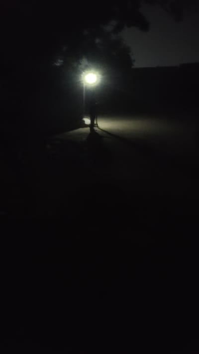 The image depicts a dimly lit scene at night in Jogana, Nigeria. A bright streetlight illuminates trees and a building's edge. Wires are visible against the building's wall. The right side of the image is dominated by darkness. No people are visible.