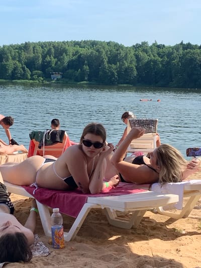 The image depicts a sunny day at a lake or reservoir, likely within the Шчомысліцкі сельскі Савет in Belarus. In the foreground, two young women are sunbathing on lounge chairs on a sandy beach. The woman on the left, wearing sunglasses, is lying on her back and looking towards the camera. The woman on the right is lying on her stomach, holding a phone or small tablet. Behind them, other people are visible in the water and on the beach, and a dense forest borders the body of water. A bottle of "PUNCH" branded beverage is on the sand next to a lounge chair.