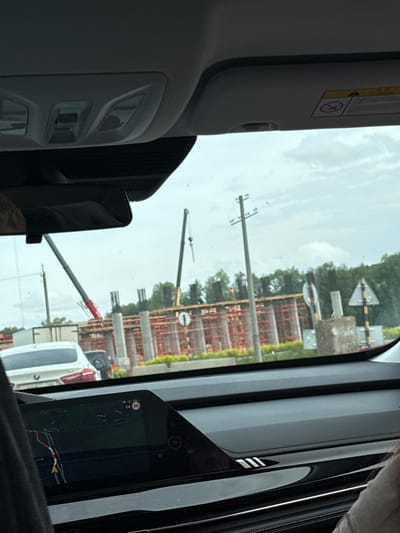 The image captures a scene from inside a vehicle, looking out the front passenger window at a construction site on an overcast day in Kabanikha, Russia.

The outdoor scene is dominated by an extensive construction site featuring orange scaffolding, concrete pillars, and rebar, suggesting the construction of a large building or bridge. Two cranes are visible, one with a red arm in the foreground and another yellow one further back. Several utility poles stand in the landscape. Two triangular road signs with black borders and white backgrounds are prominent: one with a black cross (indicating a railway crossing) and another with a vertical arrow symbol. Trees and general vegetation are visible in the background, and a white car is partially seen behind the construction.

Inside the vehicle, the sun visor is visible in the top left corner, displaying a yellow "WARNING" label with text and a graphic of a child in a car seat with a diagonal line through it, advising against placing rear-facing car seats near active airbags. On the right, a digital display shows a navigation map with road layouts, various icons, and the numbers "14 50" within a circular icon, likely indicating a speed limit. Other interior details include passenger-side air vents or controls and a side pillar with three distinct vertical white stripes. The image appears to be taken during daytime, with a cloudy sky.