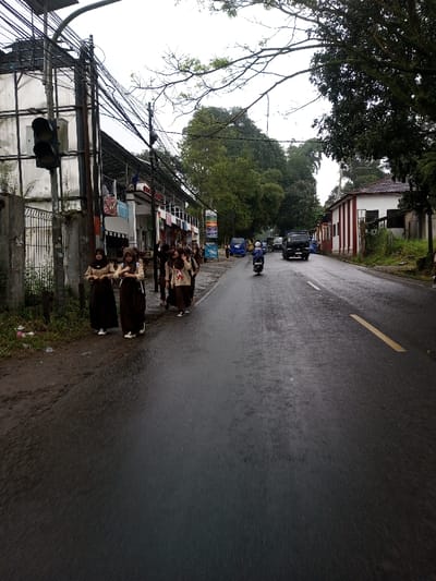 The image presents an overcast, wet street scene. On the left side of the asphalt road, a group of approximately seven to nine young females, appearing to be students, walk along a dirt shoulder. They are dressed in uniforms consisting of dark brown long skirts or trousers, light-colored long-sleeved tops, and several wear brown headscarves (hijabs). They carry bags. The road surface is visibly wet, reflecting the gray sky. A faded yellow dashed line is present on the road.
A motorcycle with a rider is positioned in the middle of the road, traveling away from the viewer. Further in the distance, a dark-colored truck and multiple blue microvans are visible.
To the left of the road, a concrete fence separates the street from a multi-story white building. A utility pole heavily laden with a dense network of black electrical and communication cables supports a black, vertically oriented traffic light with three lamps. Beyond this, a series of commercial buildings or shopfronts are visible, with clothing and other items displayed. A colorful advertising banner is also present.
On the right side of the road, a single-story white building with prominent vertical red stripes and a reddish-brown tiled roof stands. Lush green trees and dense foliage border the scene, particularly on the right and in the background. The environment is urban or semi-urban under cloudy conditions.
The visual characteristics, including the specific style of school uniforms, architecture, and vehicles, are inconsistent with the specified location of YonPhoogla, Bhutan, and are more characteristic of a Southeast Asian country like Indonesia or Malaysia.