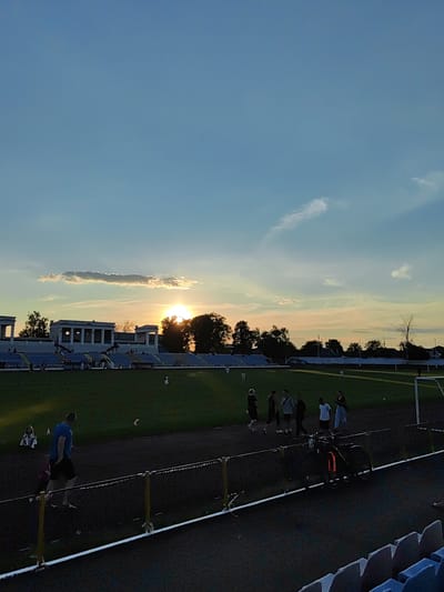 The image shows a sunset over a stadium in Korosten, Ukraine. The sky is a gradient of blue and orange, with some clouds. The sun is setting behind trees and buildings, casting long shadows across the field. There are several people in the stadium, some of whom appear to be playing or practicing on the field. There are also bleachers for spectators, some of which are occupied. A bicycle is parked near the track. The stadium appears to be well-maintained, with green grass on the field and clean bleachers. The overall impression is one of a peaceful evening in a community setting.