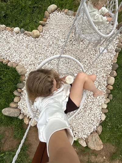 An overhead shot captures a person relaxing in a white hammock chair. The chair is suspended by thick white ropes and is positioned over a bed of white gravel and scattered larger stones. To the left of the hammock, another similar hammock chair is visible, with a child lying in it, wearing a white hooded sweatshirt.

The person in the main hammock chair, who appears to be an adult, is wearing a white long-sleeved shirt and dark shorts. They are holding a clear plastic cup with a lid and a straw, likely containing a beverage. Their legs are crossed, and their feet are bare. Their head is tilted back, and their blonde hair is spread around them.

The surrounding environment is green grass interspersed with dirt patches. The weather appears to be overcast or that the photo was taken during a time of day when the light is not direct. The overall scene suggests a casual outdoor setting, possibly a backyard or a recreational area. The emotions conveyed are relaxation and leisure.

There is no visible text in the image. The location context of Ryzhkovo, Russia, places this scene within a specific geographical area, though no visual cues within the image itself definitively point to this location.