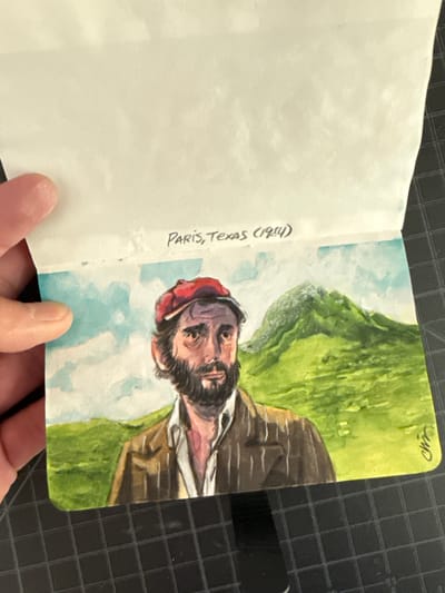 The image shows an open sketchbook. The left page is blank. The right page contains a watercolor painting of a bearded man wearing a red hat and a striped jacket, set against a backdrop of green hills and a light blue sky. The text "Paris, Texas (1984)" is written in black ink on the left page, adjacent to the painting. The sketchbook is resting on a black gridded surface. A person's hand is holding the bottom of the sketchbook. The location is irrelevant, as it is not visible in the picture.