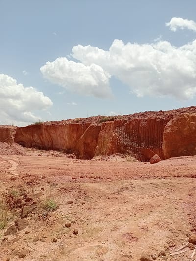 The image depicts a large, excavated area with reddish-brown earth. A steep, layered rock formation with visible vertical striations forms the backdrop, suggesting a quarry or landslide. The foreground consists of uneven, dry soil scattered with small rocks and sparse, dry vegetation. The sky above is a light blue with scattered white, puffy clouds. The scene appears to be an outdoor, undeveloped site, likely in Rayfield, Nigeria, given the context provided, possibly an area undergoing excavation or showing natural erosion. No people or specific objects beyond natural elements are visible.