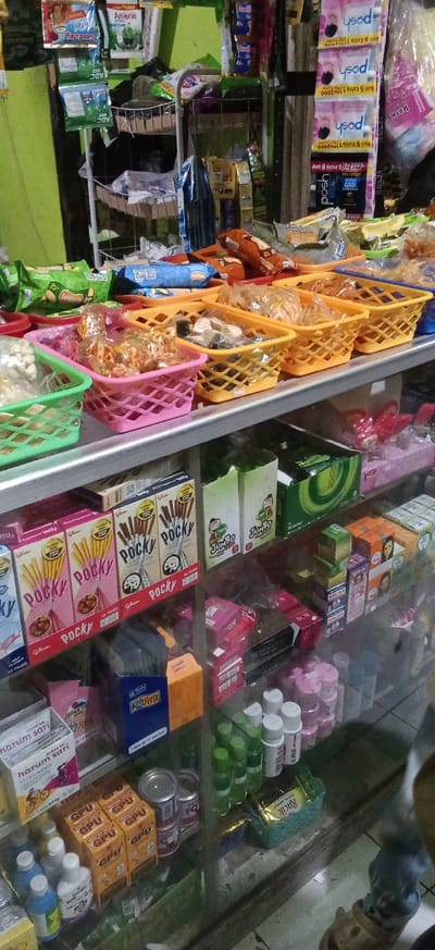 The image depicts a retail display shelf in a small store, likely a convenience store or warung, in Banjaran, Indonesia. The shelf is stocked with a variety of packaged goods, primarily snacks and personal care items.
In the foreground, several colorful plastic baskets contain assorted snacks. A pink basket holds small, individually packaged items, some appearing to be crunchy snacks. Next to it, a yellow basket contains what looks like dried crackers or biscuits. Further to the right, another yellow basket holds similar items. Green and blue packaging is visible on the shelf below.
Dominating the middle shelf are several boxes of Pocky snacks, identifiable by their distinct packaging featuring chocolate-dipped biscuit sticks. Boxes with "JUMBO" branding are also present. Other items on this shelf include various packaged foods in colorful boxes and wrappers, as well as some small green boxes.
The bottom shelf displays a mix of products. There are boxes of "HARUM SARI" products, some small cylindrical containers, and what appear to be health or personal care products in small bottles and jars. Cans and a packaged item with "Racik" branding are also visible.
The overall environment suggests a densely packed retail space, with products organized on shelves for customer access. The lighting is typical of an indoor store setting. There are no people clearly visible in the image.