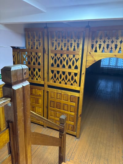 The scene depicts an interior view within a building in Bayronovka, Russia. In the immediate foreground on the left, a light-to-medium brown wooden staircase balustrade with a prominent square newel post is visible, descending towards the lower right. Across the midground, a series of large, light-brown wooden partitions function as screens or sliding doors. These partitions feature two upper tiers of intricate, openwork latticework with stylized organic patterns, and a lower tier composed of numerous small, square raised panels. They are suspended from a dark overhead track mounted to a plain white ceiling. An arched opening is present on the rightmost partition, leading into a darker recessed hallway or room. At the far end of this secondary space, a window with light-colored, possibly sheer, curtains is partially visible. The floor throughout the visible area consists of wide wooden planks, painted a worn orange-brown color, showing extensive scuffing and areas of chipped paint. No individuals or observable actions are present.