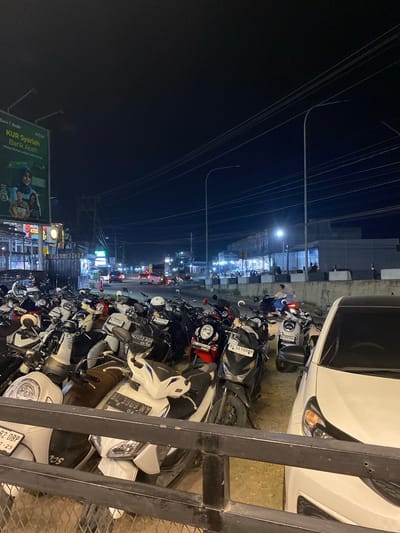 At night in Seuneubok Teungoh, Indonesia, a dense row of parked motorcycles, including many Honda scooters, dominates the foreground, stretching along the edge of a street near a white car and a metal railing. A billboard advertising "KUR Syariah Bank Aceh" is visible. Streetlights illuminate a row of shops and buildings in the distance. Power lines are strung overhead. The general setting is that of a busy urban street.