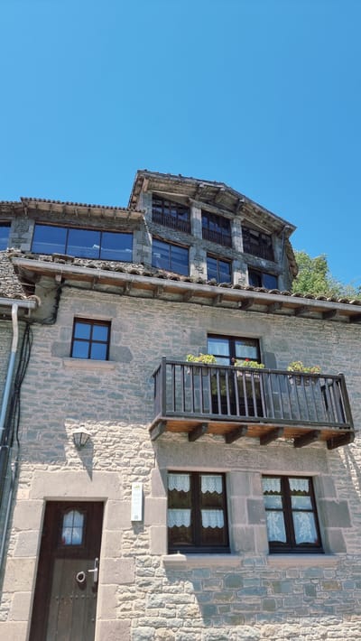 A stone building with a tiled roof stands against a clear blue sky. The building has multiple levels with various windows, some featuring white lace curtains. A wooden balcony with a railing extends from one of the upper floors, adorned with potted plants. Below the balcony, a dark wooden door with a metal handle and a small window is visible. The stone construction is evident throughout the building's exterior. A drainpipe runs vertically along the left side of the structure. The scene appears to be an outdoor shot, possibly on a sunny day, given the bright sky and shadows. The image is located in Rupit, Spain.