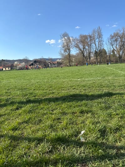 The image depicts a grassy field in Dushevo, Bulgaria, under a clear blue sky with scattered clouds. A row of leafless trees lines the edge of the field. White painted lines mark the field for sports. People are visible in the distance, near buildings at the edge of the field. Litter is visible on the grass in the foreground.