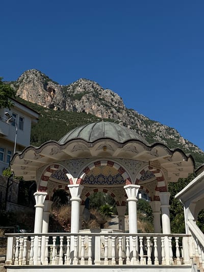 The image presents a sunlit daytime scene in Kaş, Turkey, showcasing a decorative architectural structure against a backdrop of imposing mountains and a clear blue sky.

In the foreground and midground, a beautifully adorned structure, possibly a gazebo or a public fountain (çeşme), takes center stage. It features a weathered, slightly verdigris-colored dome resting on ornate supports and white pillars. The arches and inner surfaces of the structure are decorated with intricate geometric and floral patterns in vibrant blues, reds, and gold, characteristic of traditional Ottoman or Seljuk art. On the right side, a white balustrade with finely turned spindles adds to the elegant aesthetic.

Behind this structure, a rugged mountain range dominates the background, with steep, rocky slopes and patches of green vegetation. A modern building with multiple windows is partially visible in the lower left, situated near the base of the mountains. The sky above is a deep, unblemished blue, indicating clear weather.

The bright, even lighting suggests it is mid-day or late morning. No human activity is visible, lending a tranquil atmosphere to the scene. Visible text on one of the ornate arches reads "HAYRAT," "M.ALİYEESİ," and what appears to be "UMMUOĞLU," implying the structure is a charitable work or public utility.