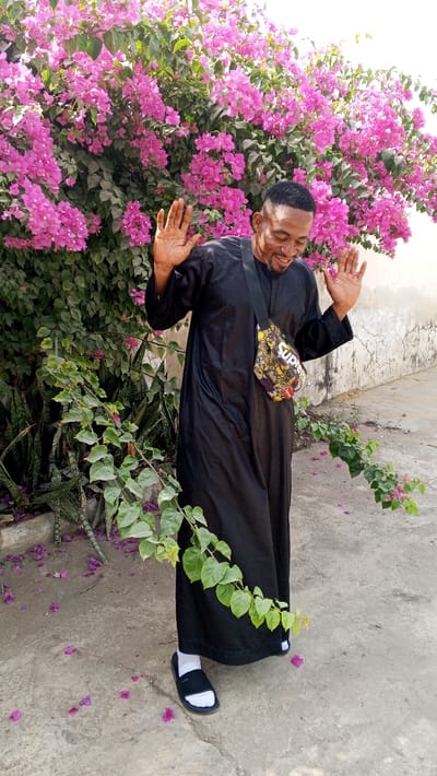 An adult male stands outdoors in Dakata, Nigeria. He has a medium skin tone, short dark hair with faded sides, and a short beard. He is smiling with his mouth open and eyes slightly closed, his head tilted. Both of his hands are raised to shoulder height, palms open and facing forward. He wears a long-sleeved black garment, white socks, and black slide sandals. A patterned fanny pack, displaying the word "SUPREME," is worn diagonally across his chest. The background features a large, dense bush covered in vibrant pink bougainvillea flowers and green leaves. Smaller green plants are visible at the base of the bush. To the right of the subject, a light-colored, weathered wall with visible cracks is present. The ground is a light grey concrete surface, scattered with fallen pink flower petals. A green vine with leaves and a few pink flowers trails across the lower foreground.