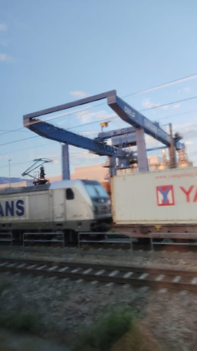 A blurred, wide shot captures a freight train moving through an industrial area, likely a rail yard, in Siezenheim, Austria. The train consists of a modern, white and silver locomotive with blue lettering that partially reads "ANS" and a series of freight cars. One car is prominently visible, carrying a large white shipping container with red and blue markings, including what appears to be a "Y" within a square. Behind the train, a large blue gantry crane, a common fixture in freight yards and ports, is partially visible, along with other industrial structures. The sky above is a pale blue with hints of clouds. The image is taken from a moving perspective, suggesting it was captured from another vehicle or train. The ground in the foreground consists of gravel and railway tracks.
