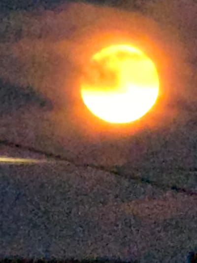 The image shows a partially obscured sun with a bright yellow and orange glow, set against a dark, textured background. The sun is located in the upper right quadrant of the image. Faint horizontal lines are visible in the lower portion of the image, suggesting the presence of power lines or similar structures. The overall scene appears to be outdoors, possibly during twilight or with atmospheric conditions affecting visibility. There are no people or identifiable objects other than the sun and implied foreground structures. The location is given as Буренка, Russia.