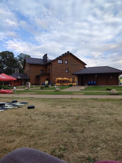 A wide shot shows a resort complex with a wooden hotel and several amenities on a cloudy day. The resort is located in Shcherbani, Ukraine. The main building is a tall, multi-story wooden structure with a dark roof. To the right of it is a smaller, single-story wooden building with a similar roof. In front of these buildings, there is a manicured lawn with a paved walkway. Several yellow umbrellas are set up over lounge chairs, suggesting an outdoor recreational area. A red inflatable lounger is visible on the left side of the image, along with a checkered game board and black game pieces on the grass. Two spherical, textured decorations are placed on the lawn. The sky is partially cloudy with patches of blue. The overall scene depicts a leisure or vacation setting.