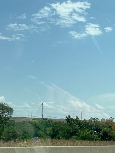 The image depicts a landscape with several wind turbines scattered across rolling hills. The sky is a pale blue with scattered white clouds, some of which appear elongated or streaky. The foreground is dominated by lush green trees and vegetation, with a blurred, light-colored horizontal line at the very bottom, possibly indicating a road or barrier. The wind turbines are tall structures with long blades, some of which are visible against the sky. The overall scene suggests an open, rural or semi-rural environment. The image does not contain any people or any identifiable elements that would suggest a specific location within Sagholasheni, Georgia, or any other city.