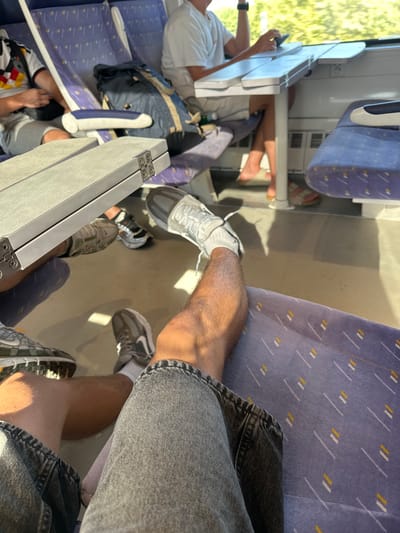 The image depicts the interior of a train carriage with a focus on passengers' legs and feet. The seats are upholstered in a blue fabric with a pattern of white and yellow lines. Two individuals are visible in the foreground, with their legs crossed and resting on the floor. One person is wearing grey denim shorts and white socks with grey and white sneakers. The other person is wearing grey shorts and grey sneakers with white socks. Another individual is partially visible in the background, seated and wearing a white t-shirt and shorts, with their hand resting on a foldable table. A navy blue backpack is placed on the seat next to them. The train carriage appears to be moving, as indicated by the blurred view of greenery outside the window. There is no specific indication of the city of Salses-le-Château, France, within the image.