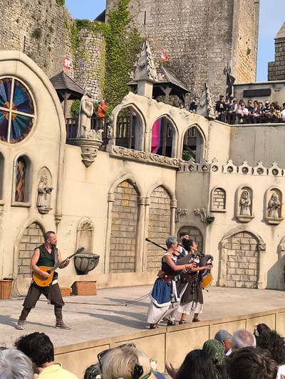 A daytime outdoor photograph captures a performance taking place on a stage in front of a castle. The stage is set with medieval-style architecture, featuring arched windows and stone textures. In the foreground, a man in a green tunic and brown trousers plays an orange lute. Behind him, two women in medieval attire are playing bagpipes. To the right of the women, another man in a brown tunic stands with his back to the camera. In the background, spectators are visible on a raised walkway in front of the castle. The castle itself is made of stone and has ivy growing on its walls. The sky is clear and blue. The scene appears to be at the Óbidos Castle in Óbidos, Portugal, a popular tourist destination known for its medieval architecture and historical events.