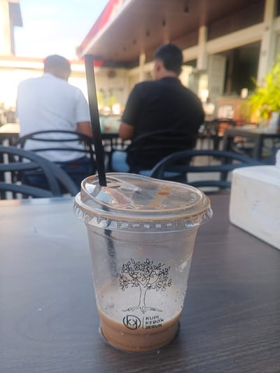 This image shows a close-up of a partially consumed iced coffee in a clear plastic cup. The cup has a black lid and a black straw. The cup is adorned with a black line drawing of a tree and the text "KUPI KEBON JERUK". The coffee itself is a light brown color with some foam on top. The cup is sitting on a dark, textured surface, likely a table.
In the background, out of focus, are two men sitting at tables, possibly at an outdoor cafe. The man on the left is wearing a white polo shirt and the man on the right is wearing a black t-shirt. There are also chairs and tables visible, as well as the facade of a building with a red awning. The sky is bright and appears to be daytime.
The image is consistent with being taken at a cafe in a populated area, possibly a city like Geulumpangsulu Timu, Indonesia, given the style of the cafe and the presence of people. The lighting suggests it is daytime, and the general ambiance points to a casual, public setting.