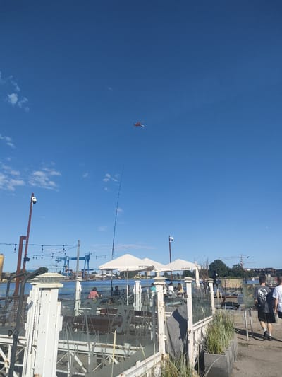 The image captures an outdoor scene on a bright, sunny day with a clear blue sky. In the distance, a kite shaped like a dragon is flying high. The foreground features a waterfront area with outdoor seating, likely a restaurant or cafe, with white umbrellas and glass barriers. Some people are seated at tables under the umbrellas, enjoying the day. A camera is mounted on a pole, and string lights are hung above the seating area.

Further in the background, a large body of water is visible, with a port or shipyard on the opposite bank. Several large blue cranes and industrial structures dominate the skyline of the port, suggesting an active working environment. The presence of these cranes and the overall industrial aesthetic hint at the location being a port city.

The overall mood is relaxed and pleasant, indicative of a leisure activity on a warm day. The clear sky and bright sunlight suggest it is daytime. The visible text "PEPSI" on the umbrellas confirms a brand presence at the venue. The context provided indicates the location is Gdańsk, Poland, which aligns with the industrial port scenery.