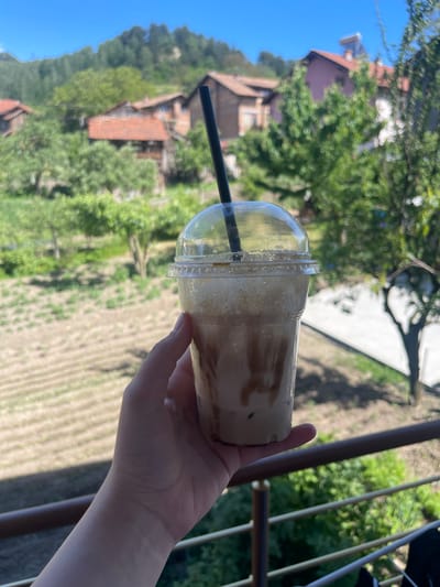 A fair-skinned hand holds a clear plastic cup of a blended coffee drink with a black straw, against a blurry backdrop showing rural Kraishte, Bulgaria. The background shows green trees and fields, residential buildings with red-tiled roofs, and a bright blue sky. A metal railing is partially visible.