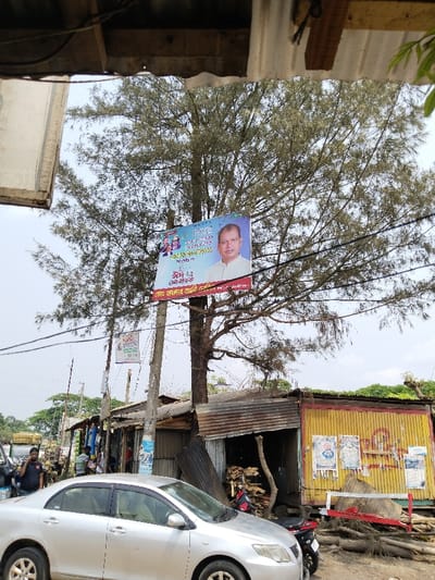 The scene is located in Pangasia, Bangladesh, at coordinates 22.705 latitude and 90.565 longitude.  A large billboard featuring a male politician, identified as "Sheikh Hasina", is mounted on a wooden pole anchored to a large tree in the foreground. The billboard is decorated with festive, colorful flowers and bears Bengali text. It hangs against a backdrop of a cloudy sky. The tree is surrounded by a collection of small shops and a single car is parked in front of one of the shops. Several people are in the scene, including a man on a motorbike and a man on foot wearing a blue shirt and a black jacket. The buildings are constructed of corrugated metal and are partially obscured by the tree. The ground is composed of dirt, debris, and scattered sticks.