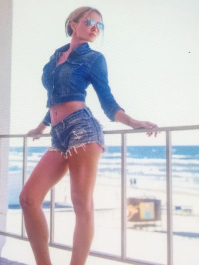 A single woman stands on a balcony, leaning forward with her hands resting on a light-colored metal railing. She has light-colored hair, pulled back, and wears reflective sunglasses. Her attire consists of a cropped, long-sleeved blue denim jacket, unbuttoned, and distressed blue denim shorts with frayed edges, exposing her midriff and navel. She is facing slightly to her right, looking out past the railing. The background reveals a wide expanse of ocean with breaking waves, meeting a sandy beach. The sky above is bright and clear, indicating daylight. In the distance on the beach, blurry figures and a rectangular, possibly structural, object with yellow and light blue elements are visible. There is no discernible visual information within the image to identify the location as Zelenyi Hai, Ukraine.