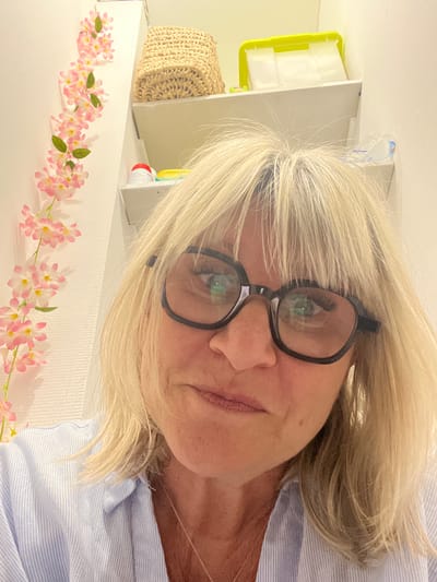 A woman with blond hair and glasses is positioned at an oblique angle in the image. She is wearing a blue and white striped shirt. In the background, there's a white shelf adorned with a pink floral garland. Other items on the shelf include a straw basket and a container with a green accent.

I am unable to confirm that this image was taken in Montélimar, France.
