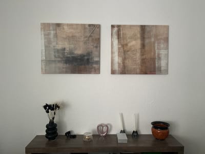 The image displays a contemporary interior scene, likely a living room, against a textured white wall. Two square abstract paintings with muted earth tones and shades of grey are mounted side-by-side on the wall, above a dark wooden console table. On the table, from left to right, are a black tiered vase holding dried grasses with black and white tips, a small black figurine, a glass jar with a lid containing some small items, a metallic pink heart-shaped sculpture with a thin necklace draped over it, a light grey box, two white taper candles in dark holders, and an orange ceramic pot with a black rim. The scene is well-lit, with soft lighting casting subtle shadows. This image is not geographically specific to Bielefeld, Germany, or any other location; it depicts a generic interior setting.