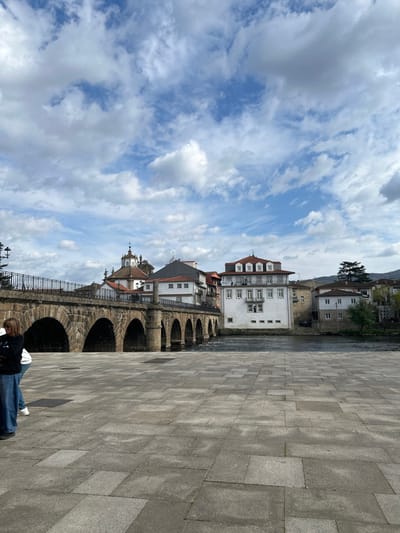 The image depicts the Roman Bridge (Ponte de Trajano) over the Tâmega River in Chaves, Portugal, viewed from a paved plaza on the left bank. The multi-arched stone bridge, constructed of light-colored masonry, features at least seven visible arches supporting a pathway topped with a dark metal railing on the left and a stone balustrade on the right.
On the left bank, immediately behind the bridge, a collection of traditional Portuguese buildings with white facades and terracotta-tiled roofs is visible. A prominent baroque-style church tower or cupola, topped with a cross, rises above these structures. Across the Tâmega River, on the right bank, several white-rendered buildings with orange-tiled roofs line the water's edge, with distant hills partially visible on the horizon to the far right.
The foreground consists of a wide, flat plaza paved with grey rectangular and square flagstones. Two individuals are present in the lower-left corner of the frame. One person, positioned on the left, wears a dark top and light blue wide-leg jeans, facing towards the left. A second person, mostly obscured by the first, is visible wearing a white top, blue jeans, and white sneakers. The sky above is partially cloudy, displaying scattered white and grey clouds against a blue background.
