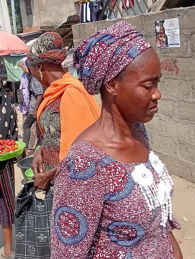 An elderly woman, with a patterned headscarf and orange shawl, sits outdoors. She wears a large patterned dress with a white lace collar and sits beside a black sheet. Her face is lined and weary. Nearby are items for sale, including tomatoes in a green container. A building with a posted flyer stands in the background, alongside a pile of clothes. The overall impression is of a scene of poverty and survival.