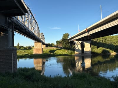 The image shows two bridges over a river in Parkany, Moldova. The bridge on the left is a metal truss bridge with concrete supports. It has a complex, industrial design. The bridge on the right is a concrete overpass with a simpler, more modern design. Both bridges appear to be in use, with the overpass possibly carrying vehicular or rail traffic. The river is calm and reflects the sky and the bridges. The banks of the river are covered in lush green grass and trees. The sky is clear and blue, with the sun casting long shadows, suggesting it is either early morning or late afternoon. There are no people visible in the image. The overall scene is one of infrastructure integrated into a natural landscape.