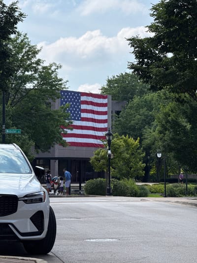 The image depicts a large American flag hanging from a building in Naperville, Illinois. In the foreground, a white SUV is parked on a street. To the left of the SUV, a street sign reads "Webster St". A lamppost stands near the sign. In front of the building, a group of people, including children, are gathered. One child appears to be wearing a football jersey. A statue of what looks like a mascot is also present. The background is filled with lush green trees and a cloudy sky. The overall scene suggests a public space on a bright, possibly summer day.
