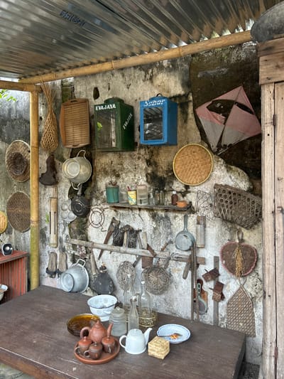 The image shows an outdoor scene with a rustic, traditional aesthetic, likely a part of a cafe or restaurant in Banguntapan, Indonesia. A weathered, rough-textured concrete wall serves as the backdrop, adorned with an assortment of vintage kitchenware and decorative items. A corrugated metal roof supported by bamboo poles covers the area.

On the wall, several items are displayed: two green and blue boxes with inscriptions, a pink and white kite, various woven baskets of different sizes and shapes, a colander, a frying pan, cooking utensils such as ladles and spatulas, and a small shelf holding bottles and containers. Below the wall, a dark wooden table occupies the foreground. On the table, a ceramic tea set with a teapot and cups, a white teapot, a bowl, and plates are arranged. To the left of the table, a red cabinet or shelf is visible.

The scene evokes a sense of nostalgia and a connection to traditional Indonesian culture, possibly as a curated display for a visitor's experience. The lighting suggests it is daytime, with natural light illuminating the scene. There are no people present in the image. The overall impression is one of an outdoor storage or display area, characterized by an eclectic collection of repurposed items.