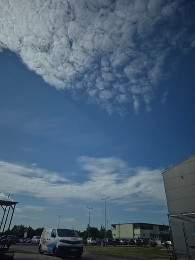 This media file captures an outdoor scene with a focus on the sky and its surrounding environment. The sky is a prominent feature, displaying a mix of fluffy, textured clouds on the left and wispy, streaky clouds on the right, against a vibrant blue backdrop. A large, modern building with a light-colored, sloped roof is visible in the upper right corner, extending from the frame.
Below the sky, a parking lot is visible, lined with several vehicles. One white van, in particular, stands out with a logo and text on its side, including what appears to be a license plate reading "WWY 807". Other cars are parked in rows. Beyond the parking lot, a grassy area and a line of trees can be seen, suggesting a natural setting adjacent to the built environment. Streetlights are also present in the parking lot.
The lighting suggests it is daytime, likely late morning or early afternoon, with clear, sunny weather indicated by the bright blue sky and distinct clouds. The scene appears to be a commercial or industrial area, possibly a business park or office building complex, given the architecture of the building and the presence of a parking lot filled with vehicles. There are no people prominently visible in the frame. The overall atmosphere is calm and ordinary, depicting a typical day in what appears to be a suburban or semi-rural location. While the image is from Pabradė, Lithuania, specific landmarks identifying this exact location are not discernible from the visual elements alone.